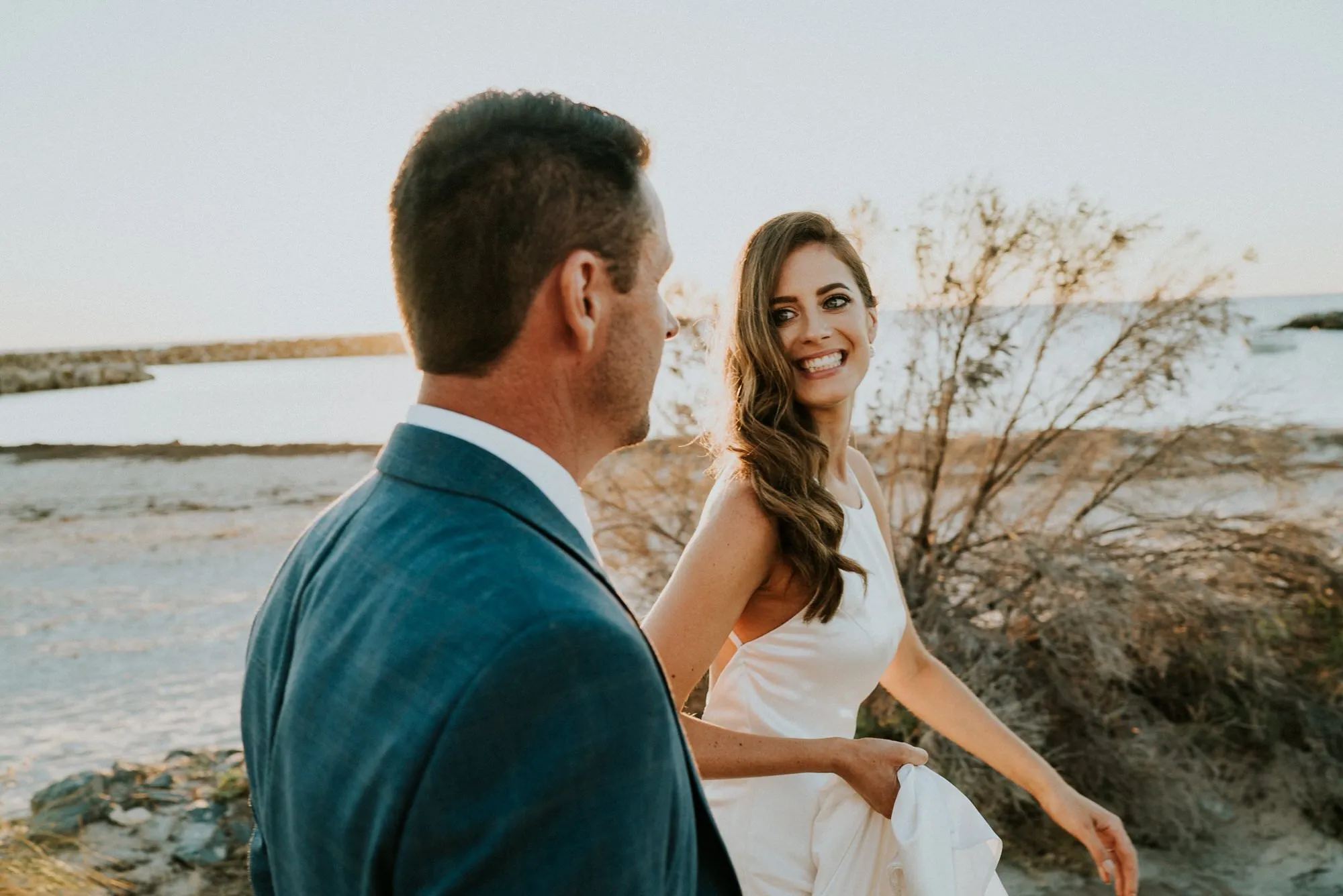 A bride glancing back and smiling at her groom on a golden-hour beach in Sydney.